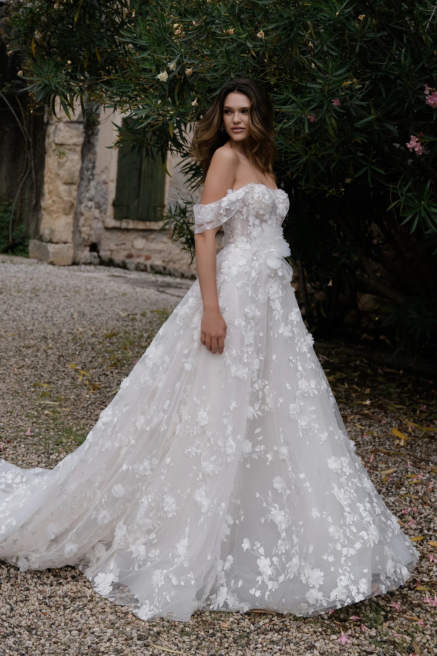 Woman in a white lace wedding dress standing outdoors with greenery and a stone building in the background.