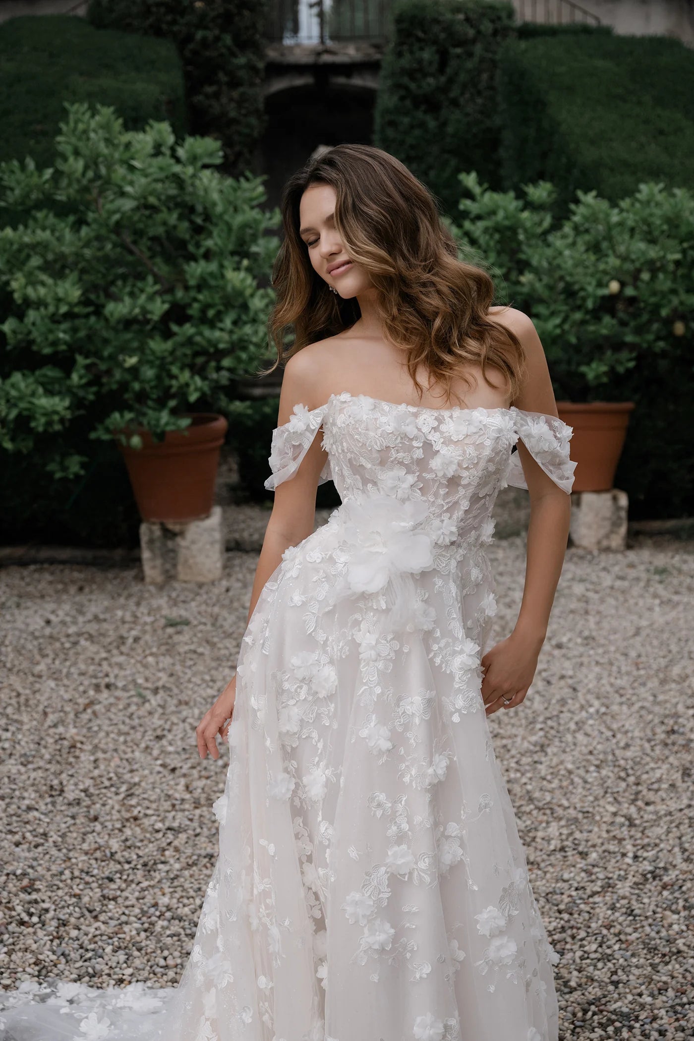 Woman in a white floral off-shoulder dress standing outdoors with greenery in the background