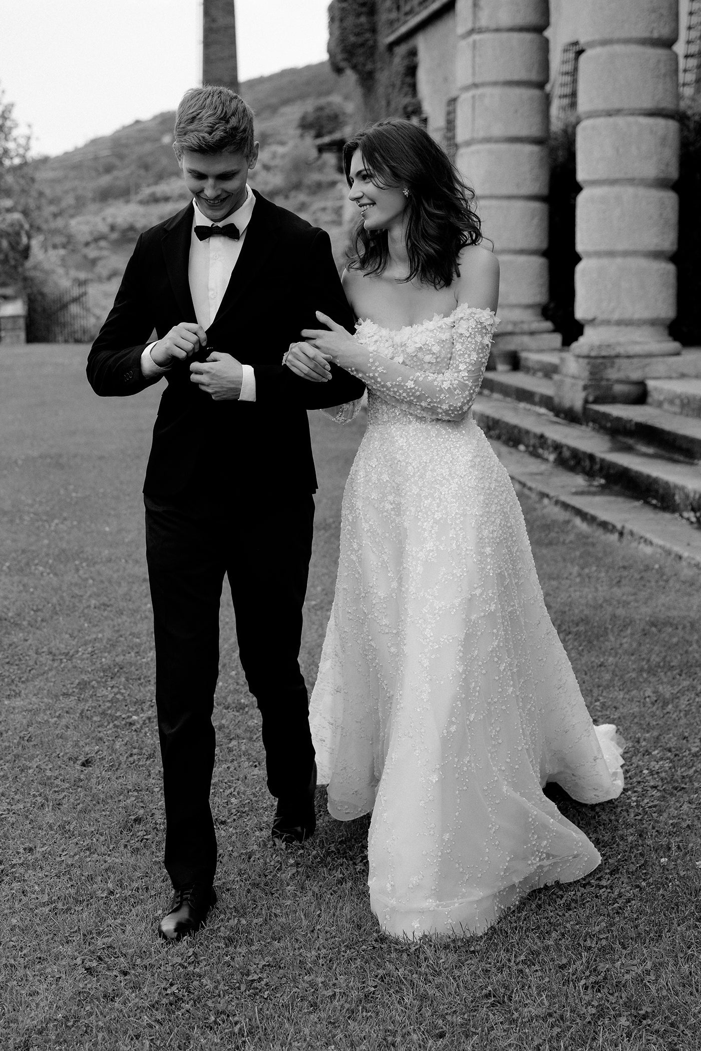 Man in a suit and woman in a sparkling dress standing outdoors with classical architecture in the background.