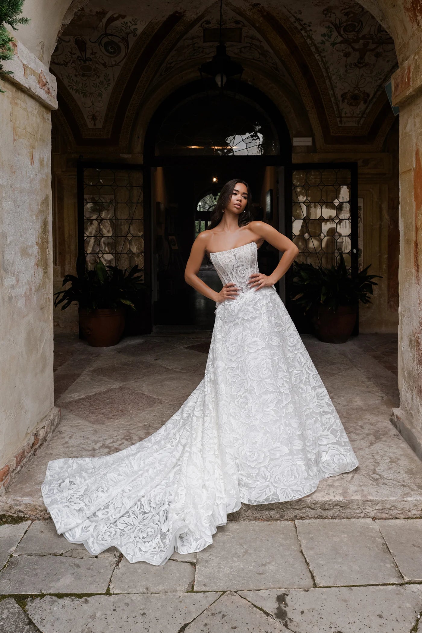 Woman in a white wedding dress standing in an elegant outdoor setting with arches and plants.