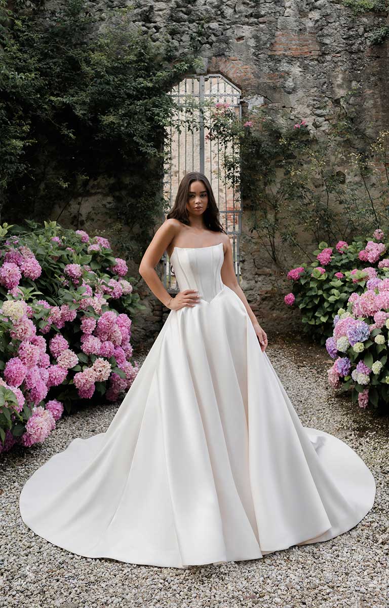 Woman in a white wedding dress standing in a garden with flowers and a stone wall.
