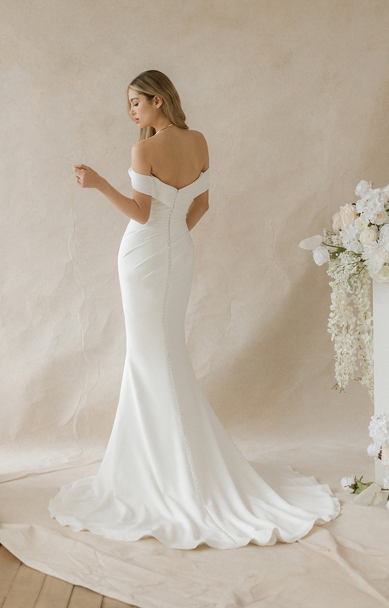 Woman in a white wedding dress standing against a neutral background with floral decorations.