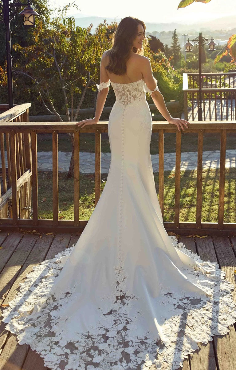 A woman wearing Aspen EK1901 by Eddy K Italia - Satin and Lace Fit-and-Flare Wedding Dress with Cat-Eye Neckline stands on a wooden deck outdoors, facing away from the camera with her hands resting on the railing. The dress has a fitted silhouette, a long lace train, and a sheer overlay near the hem.