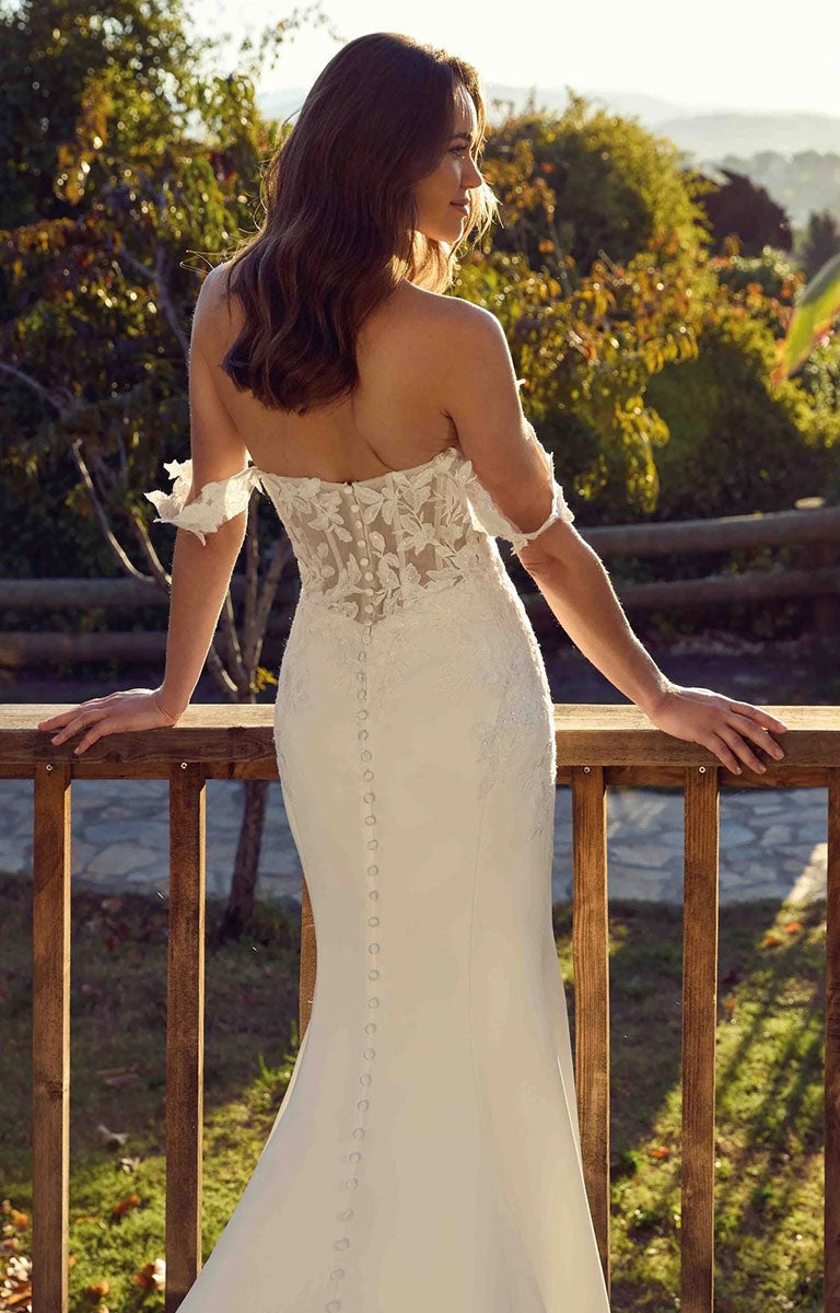 A woman wearing Aspen EK1901 by Eddy K Italia - Satin and Lace Fit-and-Flare Wedding Dress with Cat-Eye Neckline with lace detailing and a buttoned back, standing outdoors on a wooden railing with trees in the background.