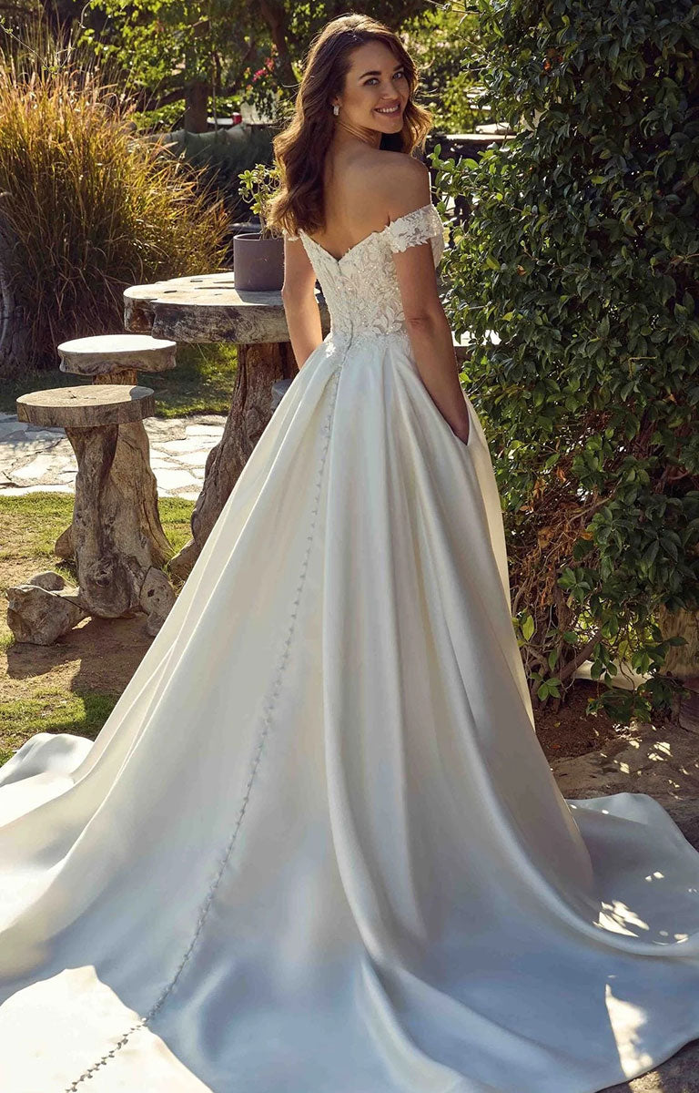 A woman wearing Opalite EK1921 by Eddy K Italia - Mikado Ballgown Wedding Dress with Chantilly Lace with a lace bodice and a long satin train, photographed outdoors in a garden setting.