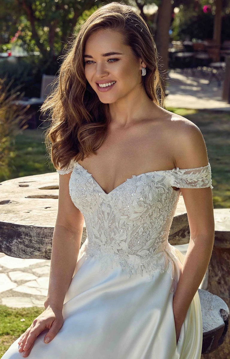 A woman wearing Opalite EK1921 by Eddy K Italia - Mikado Ballgown Wedding Dress with Chantilly Lace with a heavily embellished lace corset bodice, sitting outdoors on a rustic stone bench.