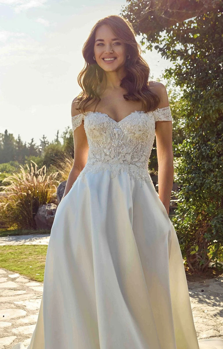A woman wearing Opalite EK1921 by Eddy K Italia - Mikado Ballgown Wedding Dress with Chantilly Lace with a lace-embellished sweetheart bodice and a flowing satin skirt, standing outdoors in a garden setting.