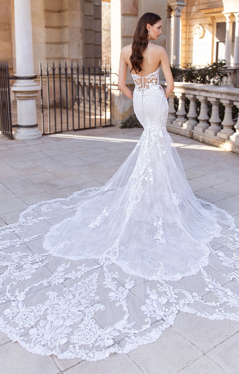Woman wearing Sibylla by ÉLYSÉE - Floral Lace Mermaid Wedding Dress with Illusion Back and Sculpted Train in an outdoor setting.