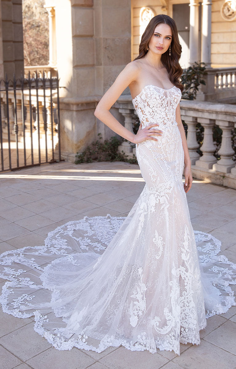 Woman wearing Sibylla by ÉLYSÉE - Floral Lace Mermaid Wedding Dress with Illusion Back and Sculpted Train in an outdoor setting.