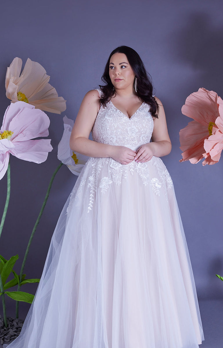 Woman wearing Amelia by Eve’s Collection - Strapped Ballgown Wedding Dress with Beaded Lace and Ruched Tulle with a lilac wall backdrop and oversized pastel colour paper flowers surrounding her.