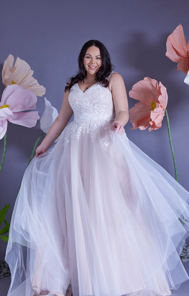 Woman wearing Amelia by Eve’s Collection - Strapped Ballgown Wedding Dress with Beaded Lace and Ruched Tulle with a lilac wall backdrop and oversized pastel colour paper flowers surrounding her.