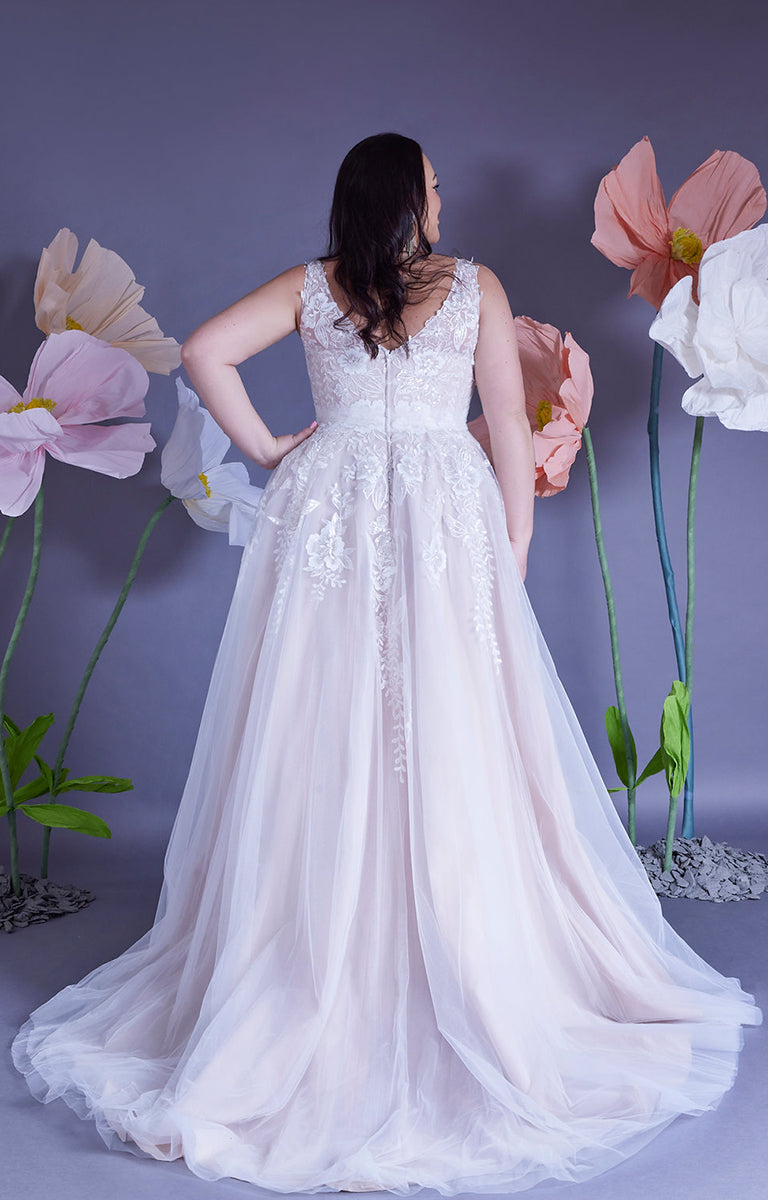 Back of a woman wearing Amelia by Eve’s Collection - Strapped Ballgown Wedding Dress with Beaded Lace and Ruched Tulle with a lilac wall backdrop and oversized pastel colour paper flowers surrounding her.