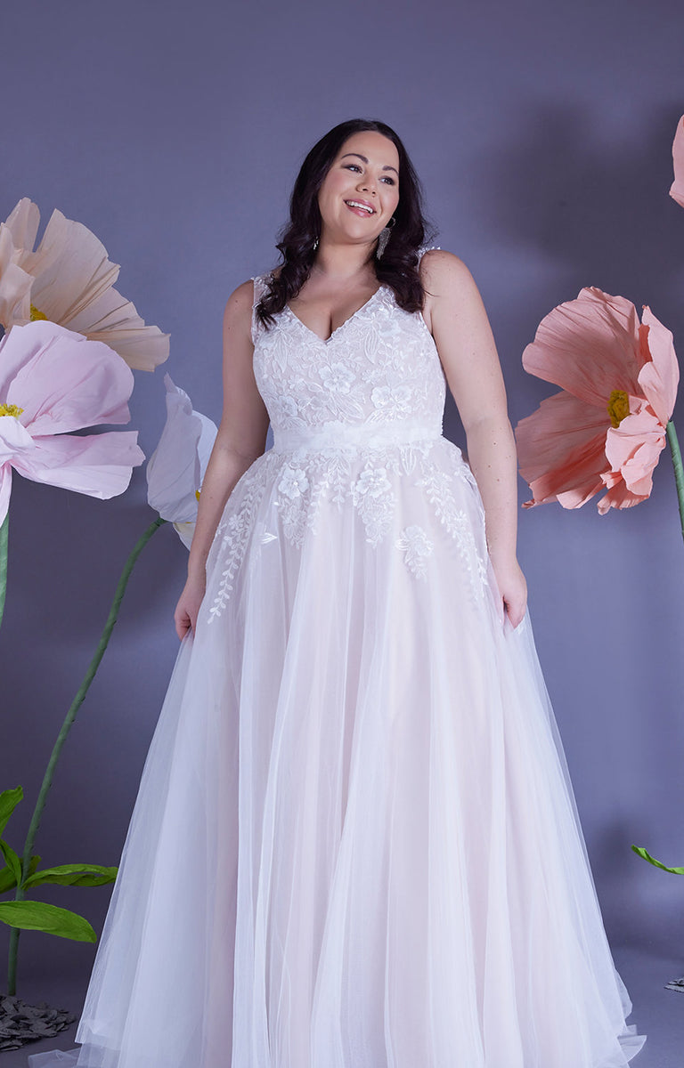 Woman wearing Amelia by Eve’s Collection - Strapped Ballgown Wedding Dress with Beaded Lace and Ruched Tulle with a lilac wall backdrop and oversized pastel colour paper flowers surrounding her.