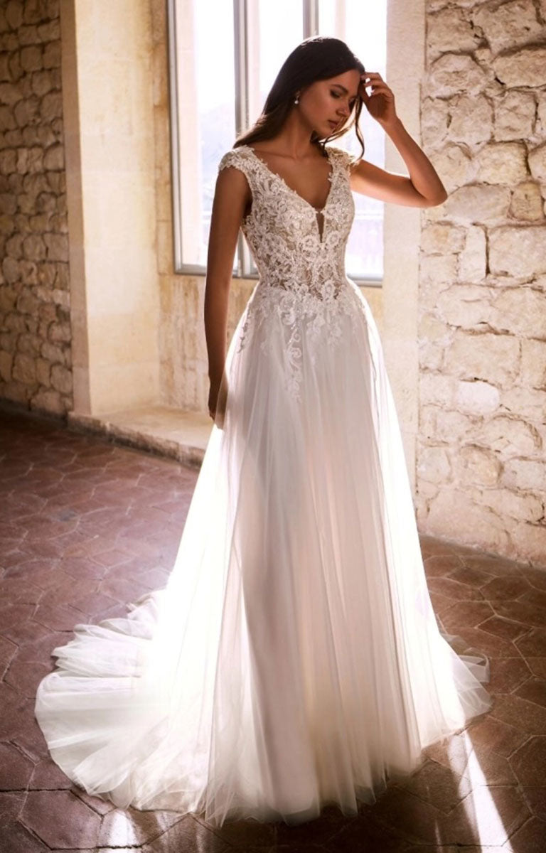 A woman wearing Gemmah by Étoile Bridal - Beaded Lace A-Line Wedding Dress with V-Neckline and Royal Train stands in a sunlit stone interior with a large window behind her.