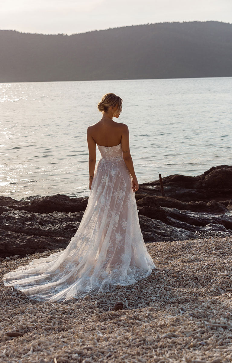 Woman in Janae by Madi Lane - A-Line Wedding Dress with Detachable Sleeves and Illusion Train standing on a rocky shoreline with water and mountains in the background.