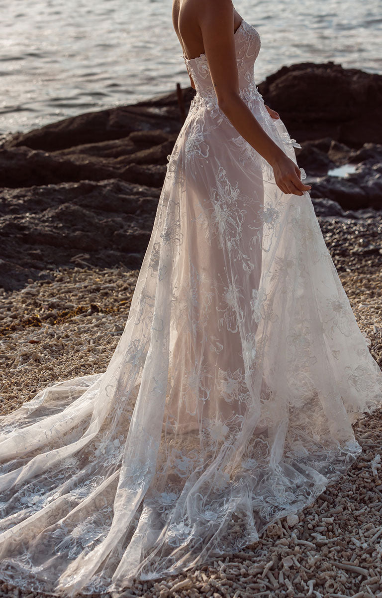 Woman in Janae by Madi Lane - A-Line Wedding Dress with Detachable Sleeves and Illusion Train stood on a beach with the sea in the background.