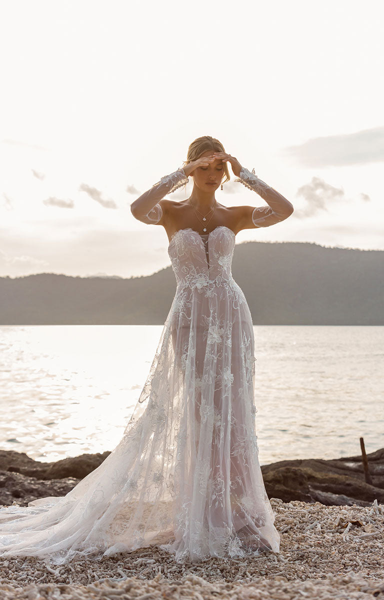 Woman in Janae by Madi Lane - A-Line Wedding Dress with Detachable Sleeves and Illusion Train standing on a beach with mountains in the background
