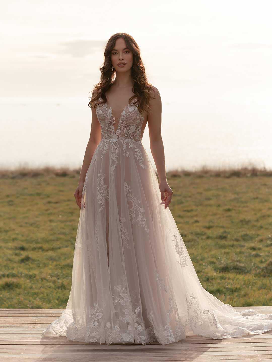 Woman in a lace wedding dress standing on a wooden platform with a grassy field and trees in the background.