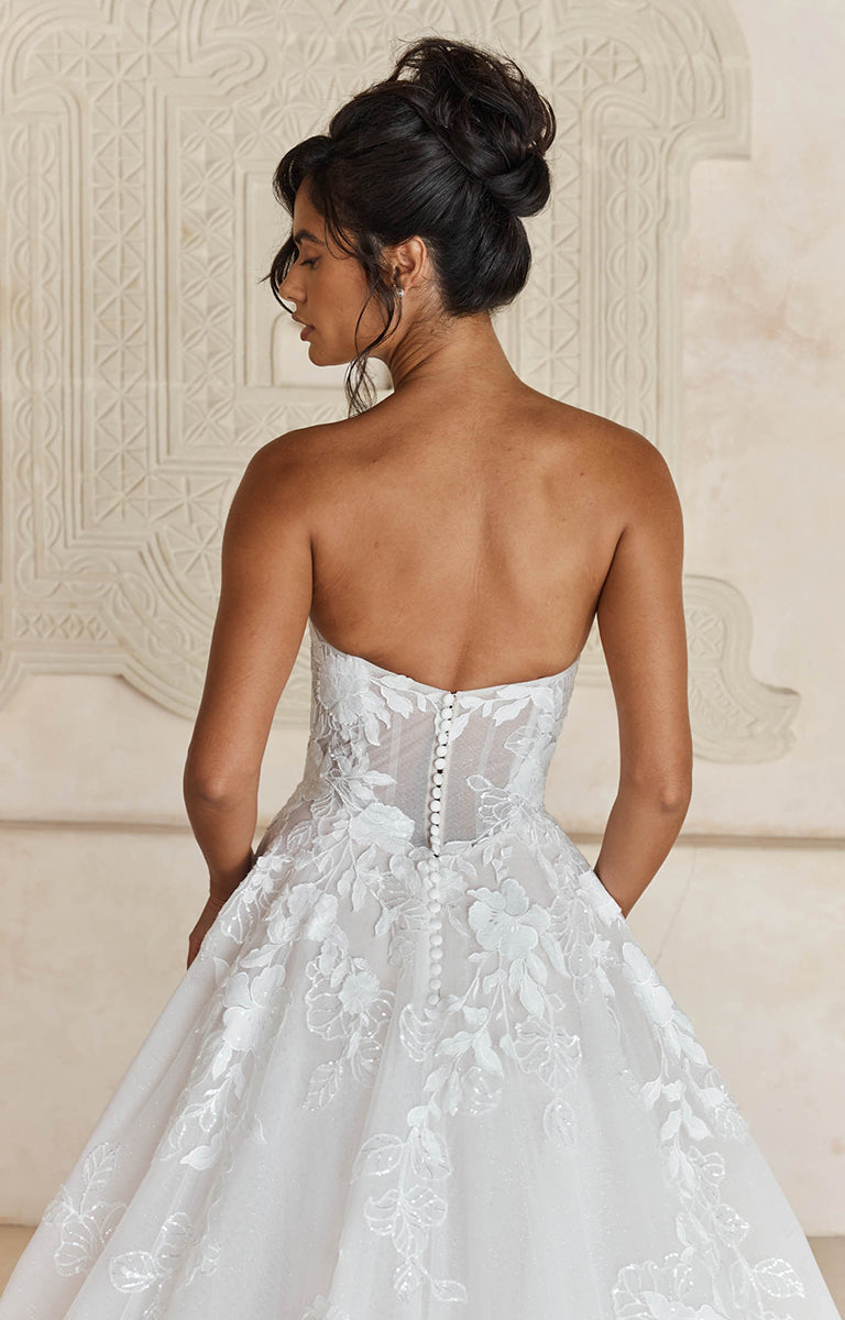 Woman wearing Liana by Madi Lane, a white wedding dress with intricate detailing, standing against an ornate wall.