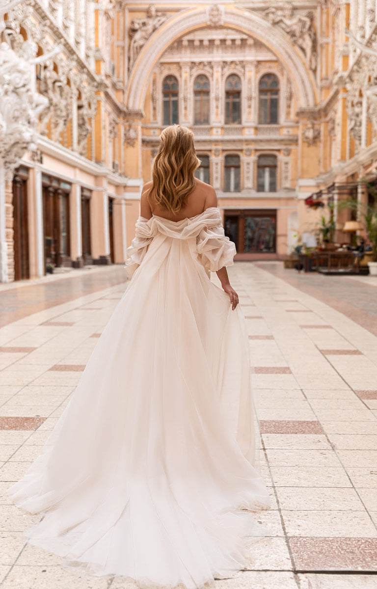 Woman in a white off-shoulder dress walking in an elegant outdoor setting with classical architecture.