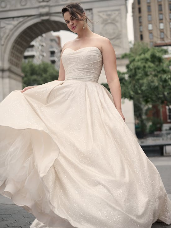 Woman in Anniston by Maggie Sottero - a strapless wedding dress standing in an urban setting with a building and archway in the background.