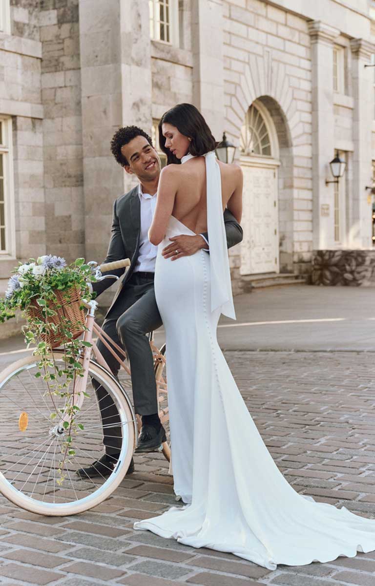 Man and woman in wedding attire standing next to a bicycle with flowers, in front of a building.