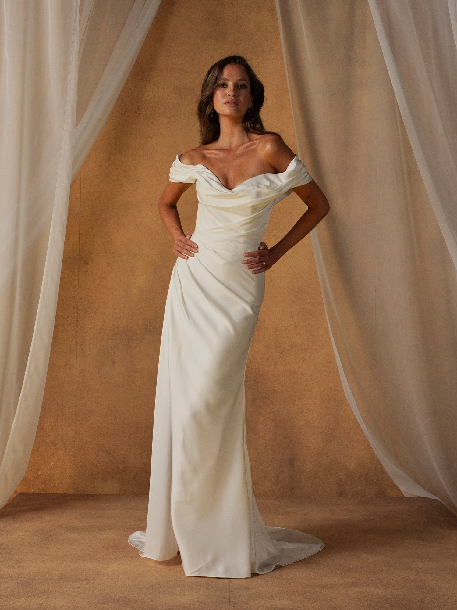 Woman in Nelson by Madi Lane - Off-the-Shoulder Fit and Flare Wedding Dress standing against a beige curtain backdrop