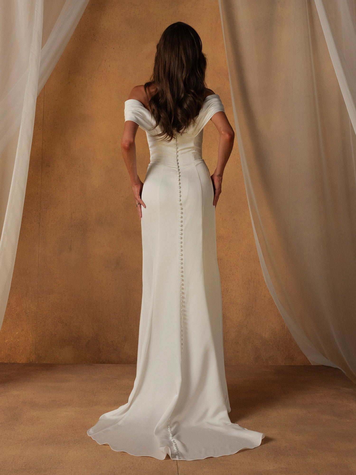 Woman wearing Nelson by Madi Lane - Off-the-Shoulder Fit and Flare Wedding Dress with button detailing, standing against a beige curtain.