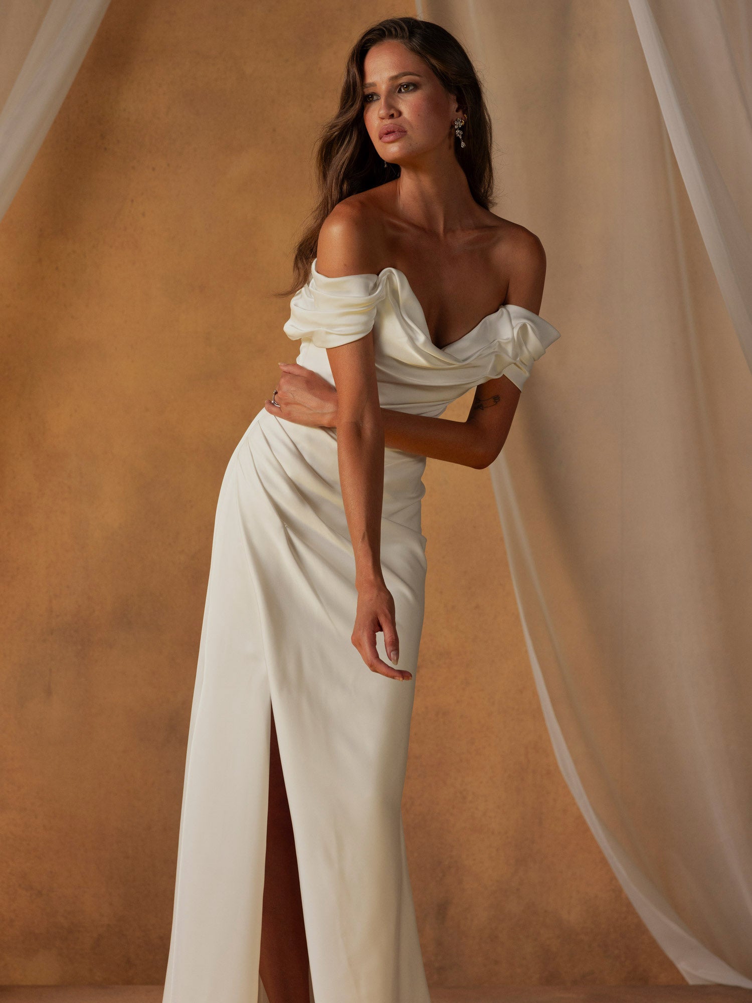 Woman in Nelson by Madi Lane - Off-the-Shoulder Fit and Flare Wedding Dress featuring a slit against a beige curtain.