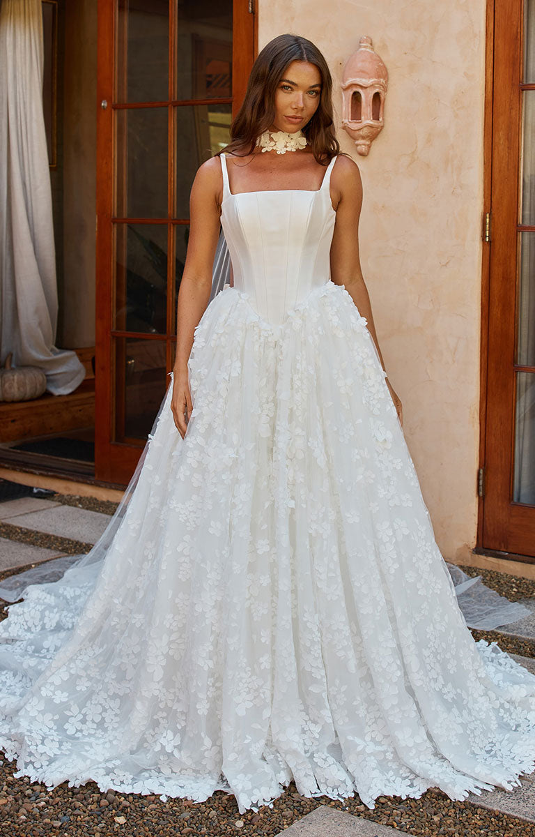 A woman modeling Orient by Madi Lane - Mikado and 3D Lace A-Line Wedding Dress with Corset Bodice and Detachable Choker with a fitted bodice, floral lace overlay, and a full voluminous skirt with a long train, standing outdoors in front of a stucco wall and wooden doors.