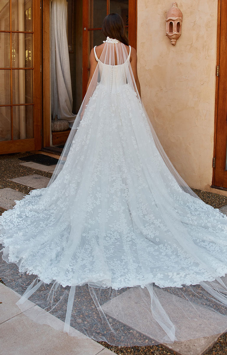 A woman viewed from behind wearing Orient by Madi Lane - Mikado and 3D Lace A-Line Wedding Dress with Corset Bodice and Detachable Choker with a long sheer tulle train and veil, standing outdoors in front of a beige stucco wall and wooden doors.