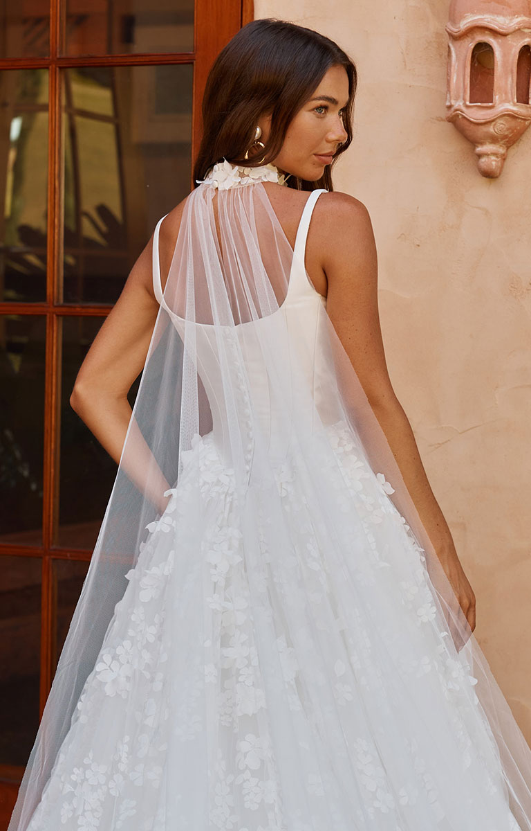 A woman wearing Orient by Madi Lane - Mikado and 3D Lace A-Line Wedding Dress with Corset Bodice and Detachable Choker with a sheer tulle back and a long matching veil, photographed from behind in an outdoor setting with a warm beige wall and window in the background.