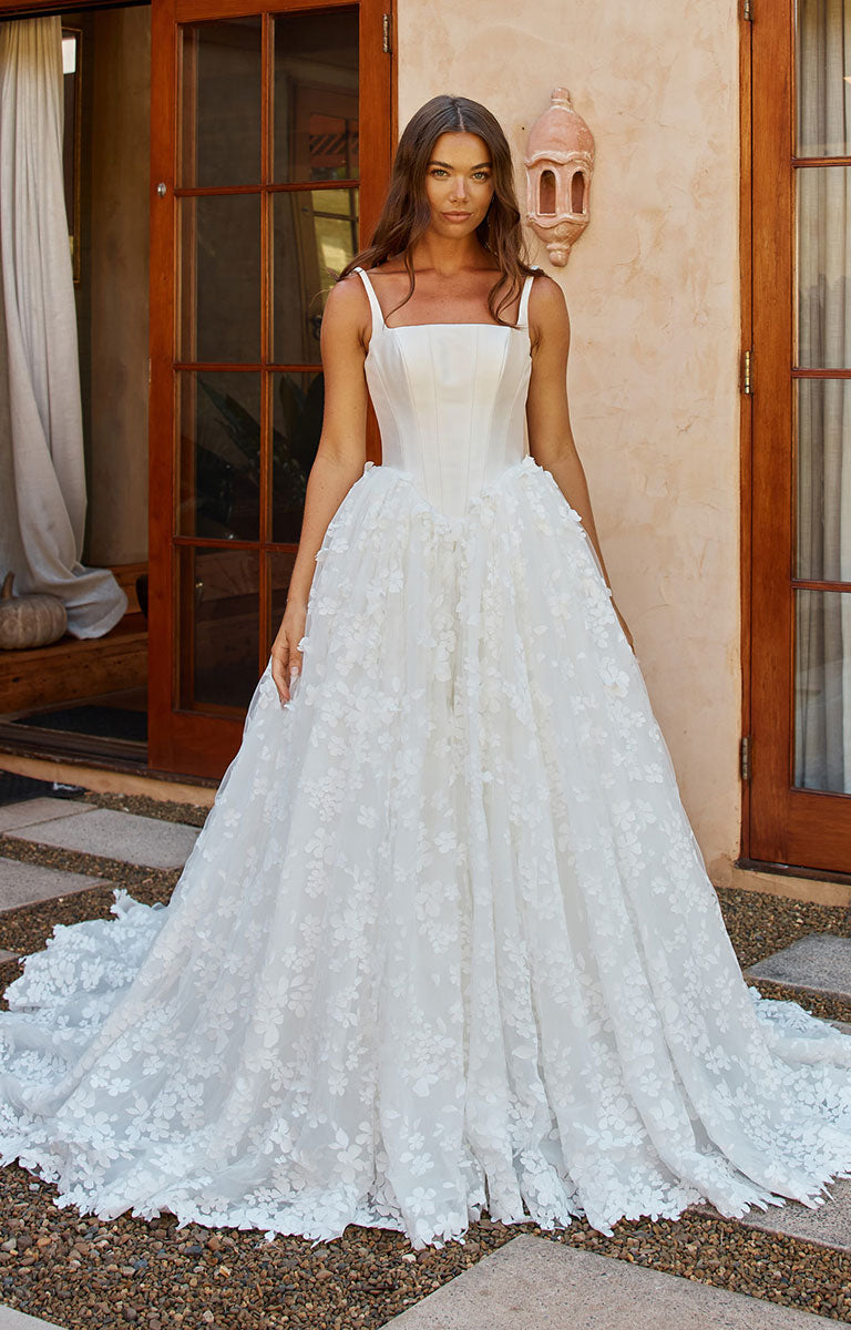 A woman wearing Orient by Madi Lane - Mikado and 3D Lace A-Line Wedding Dress with Corset Bodice and Detachable Choker with a fitted bodice and a voluminous floral lace skirt, standing outdoors in front of a beige stucco wall and wooden doors.