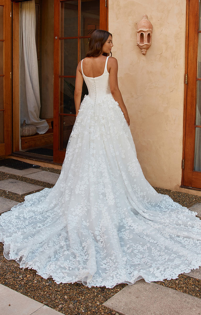 A woman wearing Orient by Madi Lane - Mikado and 3D Lace A-Line Wedding Dress with Corset Bodice and Detachable Choker is photographed from the back in an outdoor setting with a stucco wall, wooden doors, and stone pavement. The dress has thin straps, a fitted backless bodice, and a full lace train.