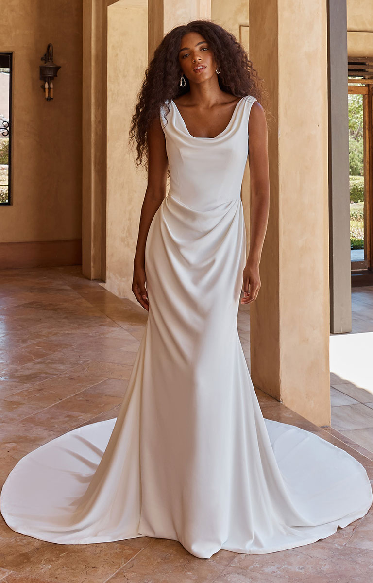 A woman modeling Ollie by Madi Lane - Draped French Crepe Fit-and-Flare Wedding Dress with Cowl Neckline with a draped satin-like finish and a full train, standing in a warm-toned architectural interior with stone columns and tiled floors.