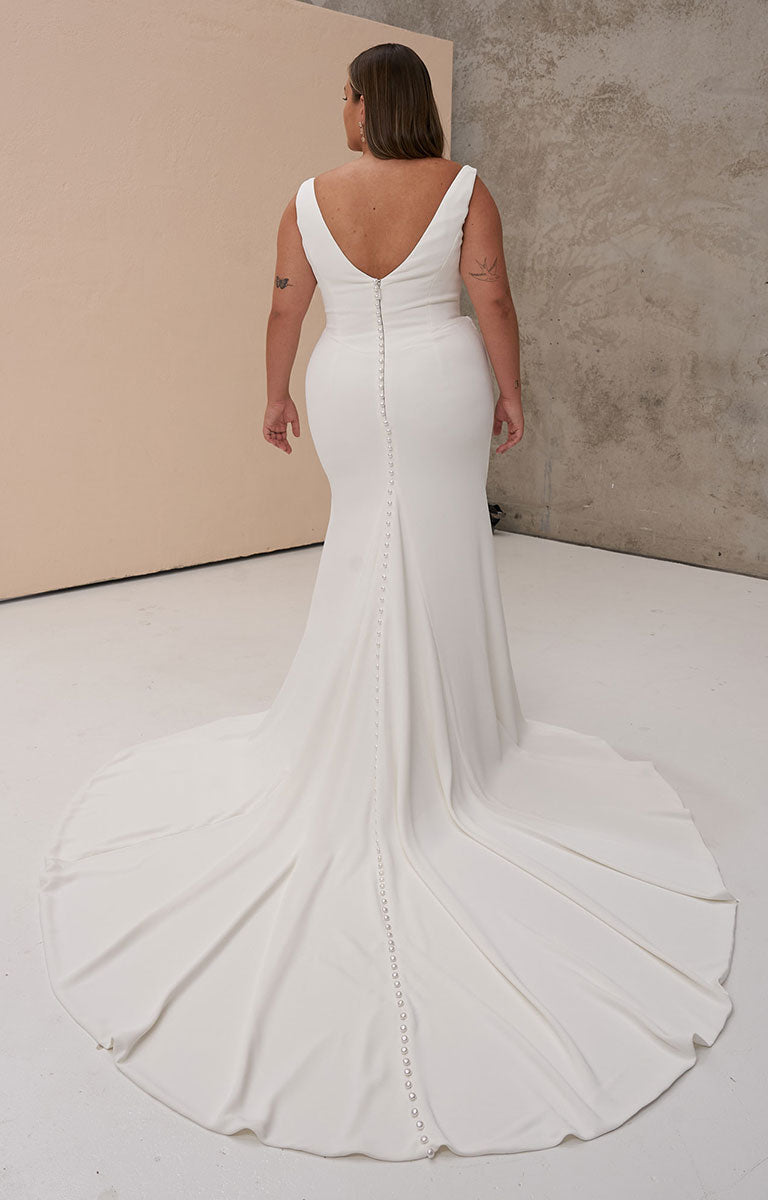 A woman stands with her back to the camera wearing Ollie by Madi Lane - Draped French Crepe Fit-and-Flare Wedding Dress with Cowl Neckline with a deep V back, center back bead detailing, and a long train in a studio with a beige backdrop and concrete wall.