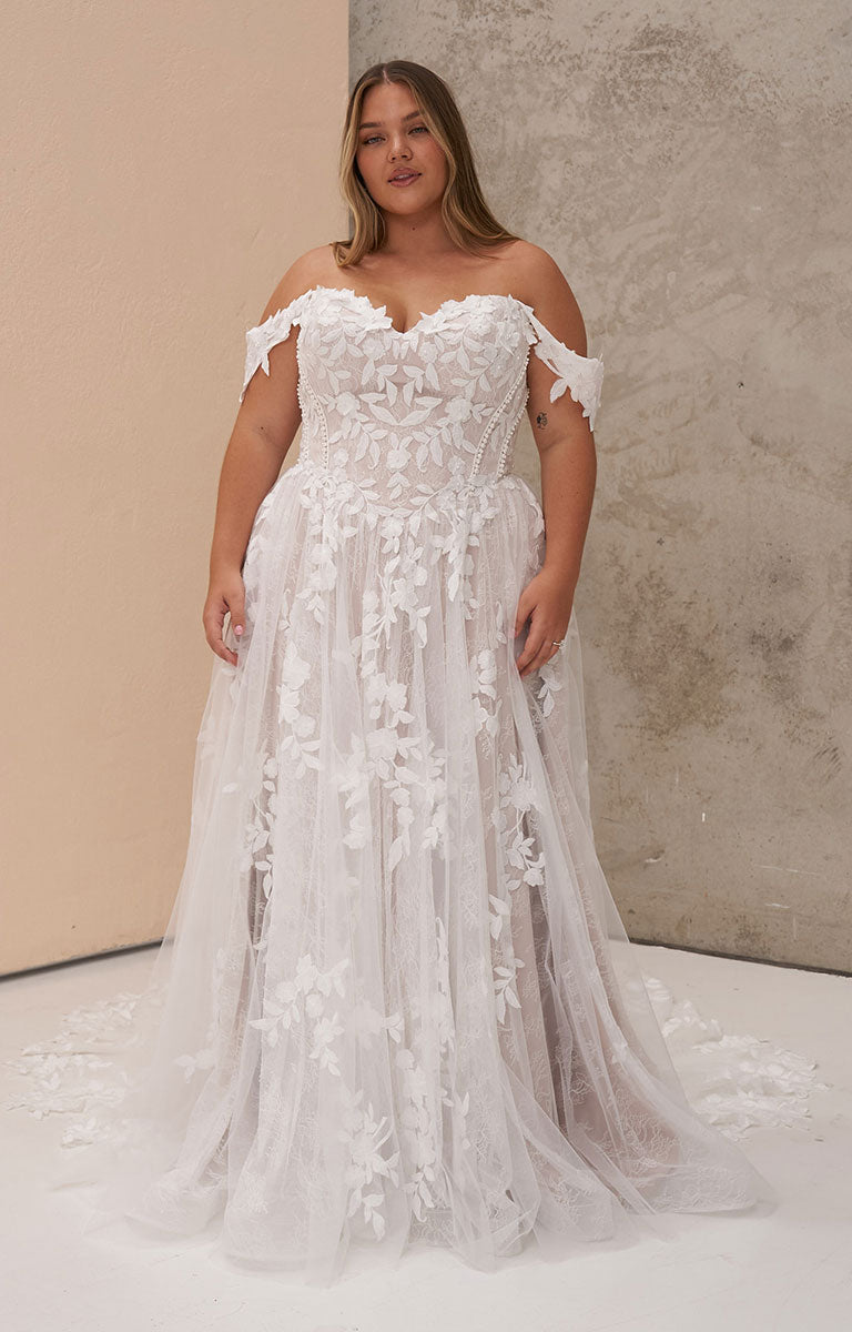 A woman modeling Osha by Madi Lane - Romantic Chantilly Lace A-Line Wedding Dress with 3D Floral Details with intricate floral lace applique and a layered tulle skirt, standing against a split beige and textured gray studio backdrop.
