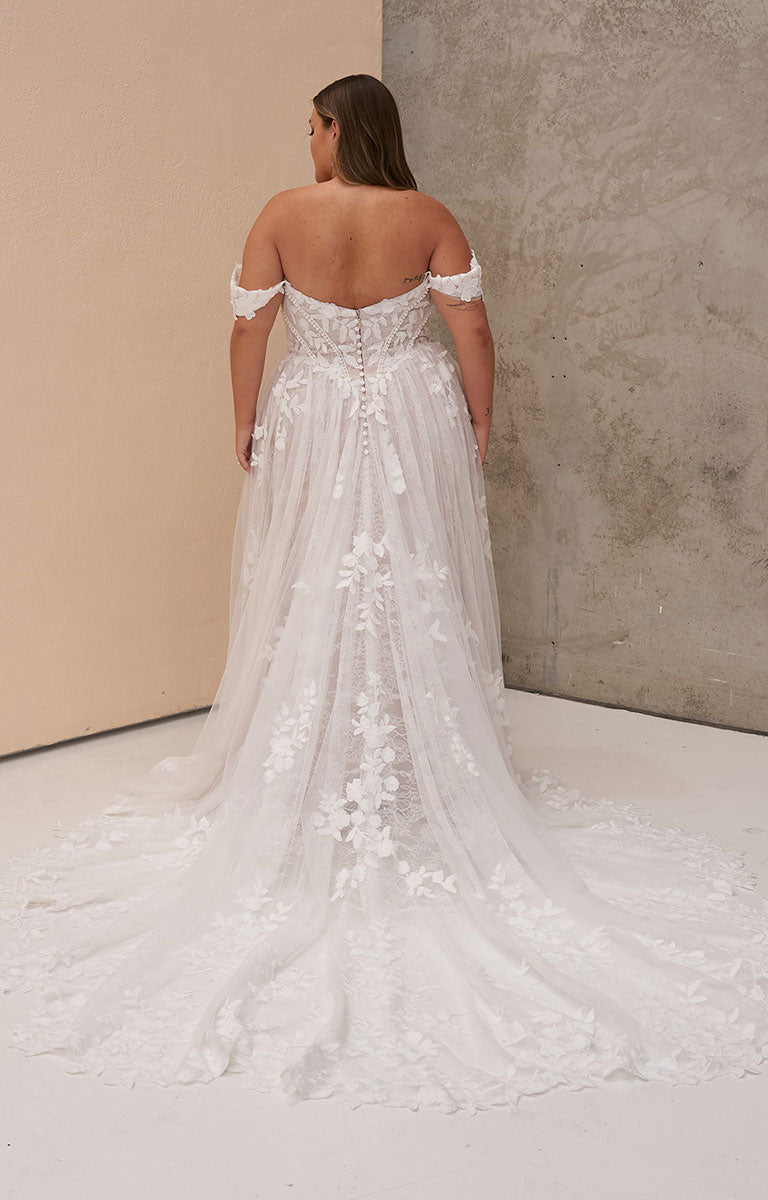 A woman viewed from behind wearing Osha by Madi Lane - Romantic Chantilly Lace A-Line Wedding Dress with 3D Floral Details with a long train, standing in a studio with a beige wall and textured gray wall background.