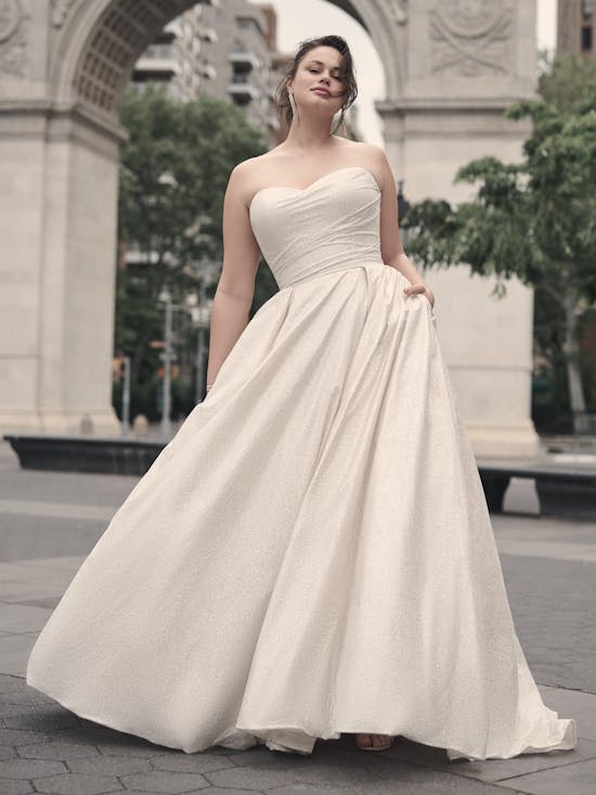 Woman wearing Anniston by Maggie Sottero - a strapless white wedding dress standing in an urban setting with a large archway in the background.