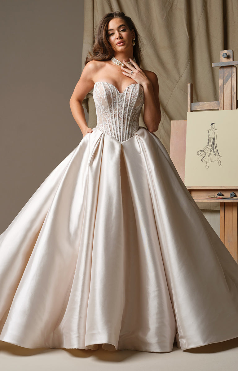 Woman wearing Ashland by Maggie Sottero - Strapless Mikado V-Neckline Ballgown Wedding Dress with a detailed bodice and flowing skirt, standing in front of an artist's easel with a drawing of a dress.