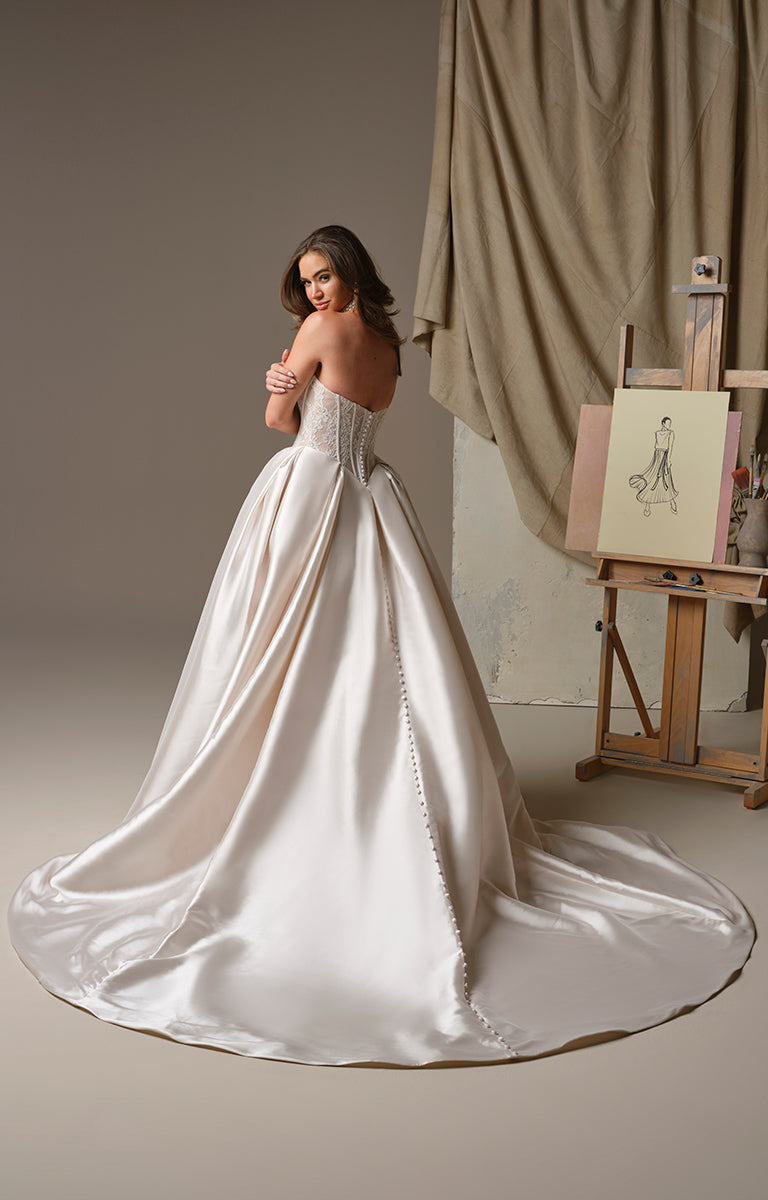 Woman in Ashland by Maggie Sottero - Strapless Mikado V-Neckline Ballgown Wedding Dress standing in a studio with an easel and painting in the background.