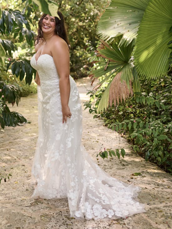 Woman in Demetria by Maggie Sottero - a white wedding dress standing in a tropical setting with lush greenery.
