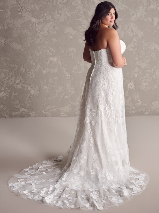 Woman wearing Demetria by Maggie Sottero - a white lace wedding dress with a long train against a neutral background