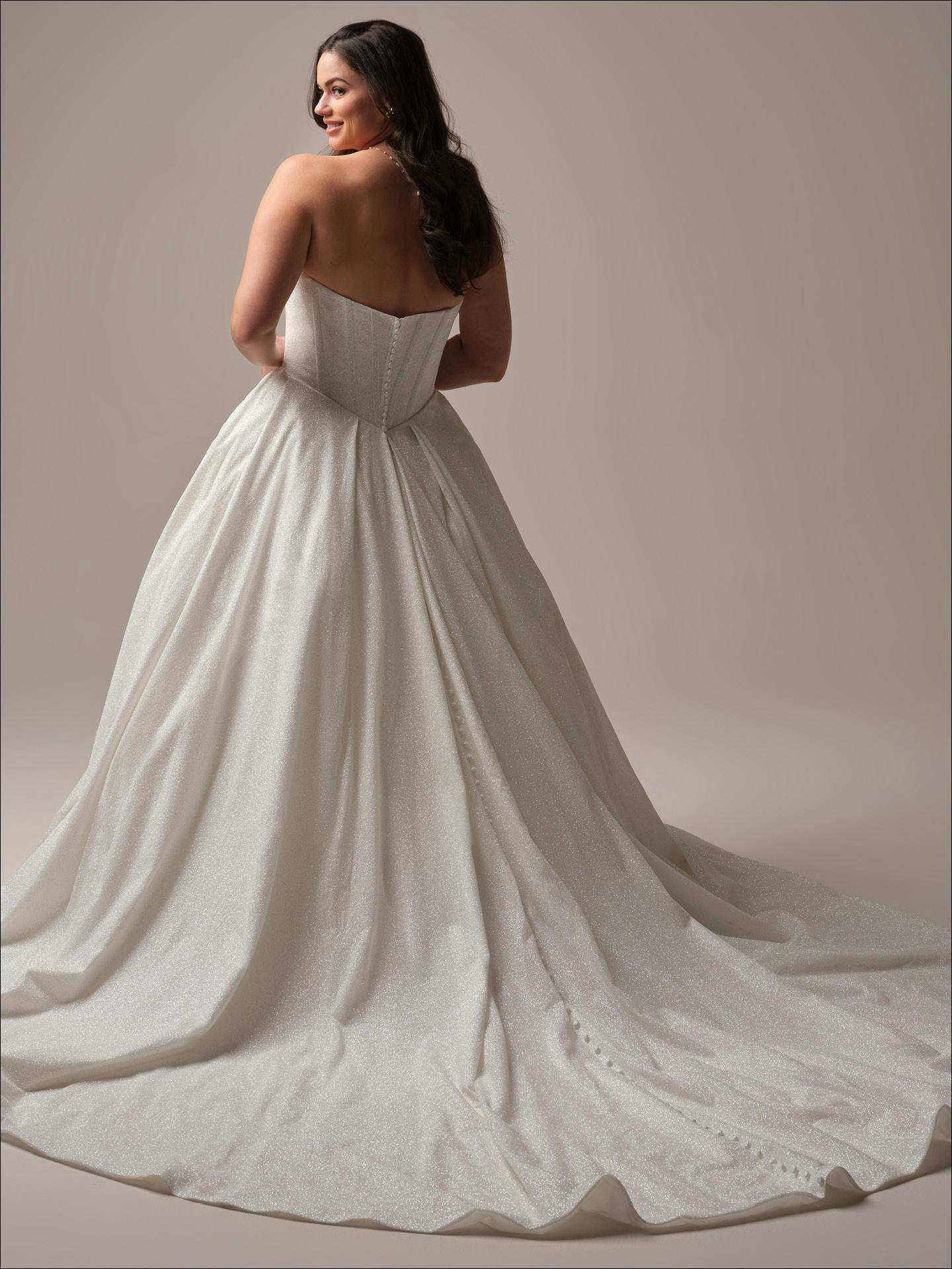 Woman wearing Derrick Royale by Maggie Sottero - a white wedding dress with a long train on a plain background
