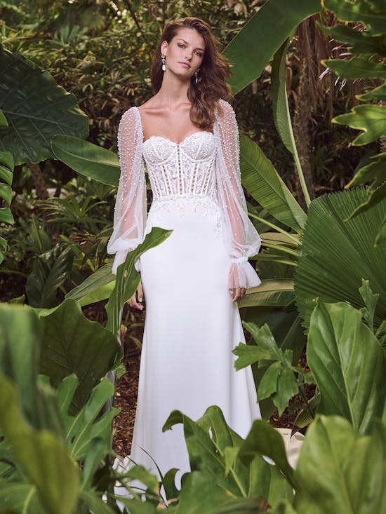 Woman in a white wedding dress with a sheer overlay standing among tropical plants.