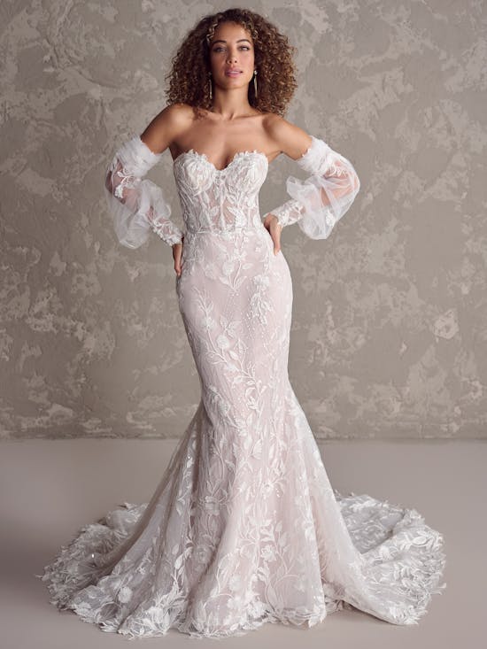 Woman wearing Fairchild by Maggie Sottero - a white lace wedding dress with long train against a textured gray background