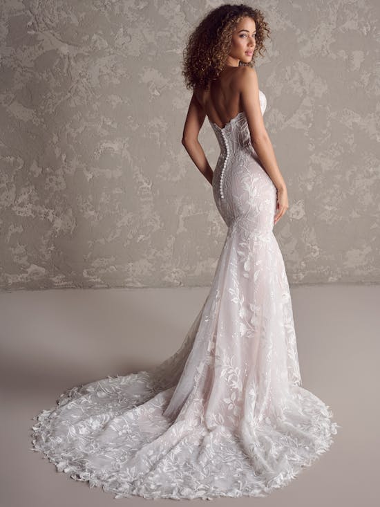 Woman wearing Fairchild by Maggie Sottero - a white lace wedding dress with a long train against a textured wall.