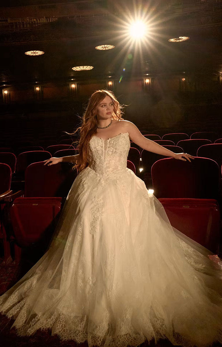 A woman in Hadley by Maggie Sottero - Beaded Lace Cat Eye A-Line Wedding Dress with Illusion Bodice stands in a dimly lit theater with red seats, illuminated by a bright overhead spotlight that creates lens flare.