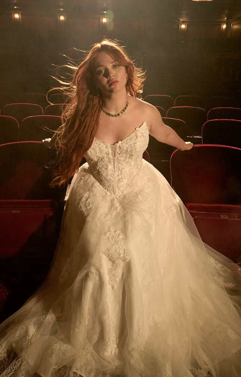 A woman with long red hair wearing Hadley by Maggie Sottero - Beaded Lace Cat Eye A-Line Wedding Dress with Illusion Bodice stands in a dimly lit theater with red seats in the background.