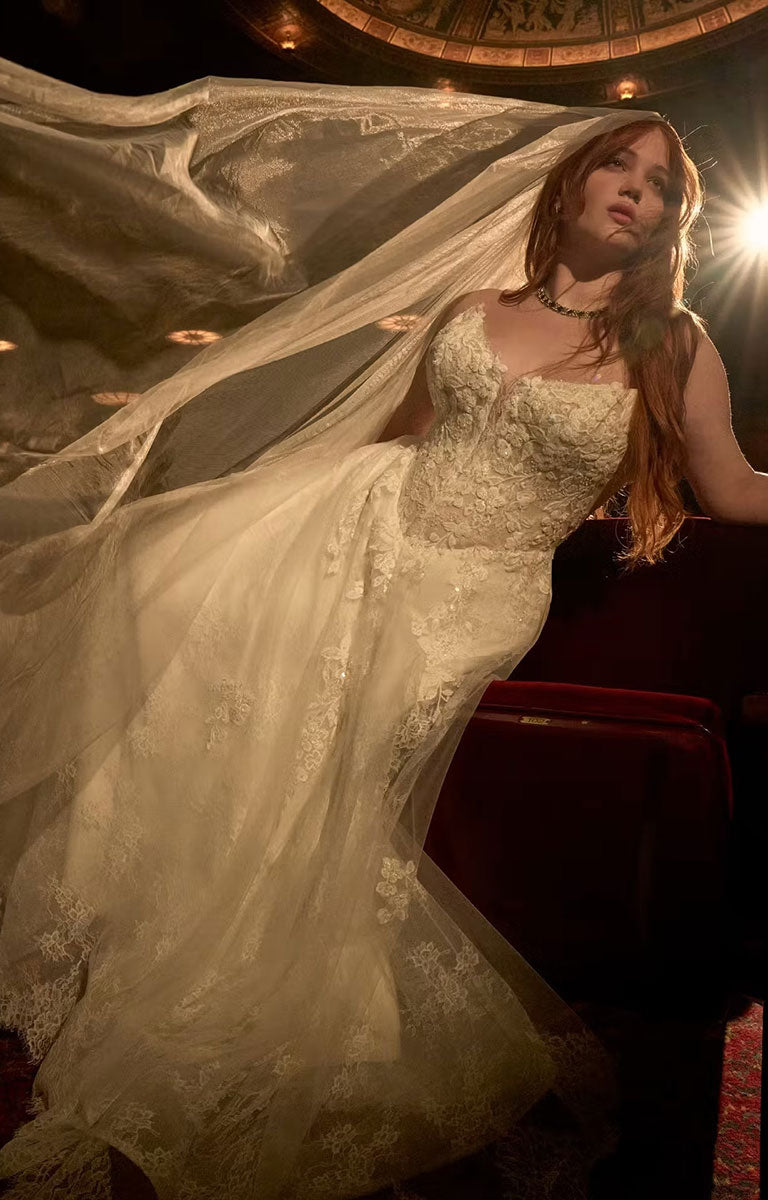 A woman wearing Hadley by Maggie Sottero - Beaded Lace Cat Eye A-Line Wedding Dress with Illusion Bodice with a long sheer veil, posed indoors with dramatic lighting and a dark red upholstered chair behind her.